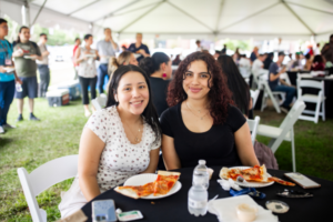 two students sitting together at a table outside smiling with plates of pizza in front of them